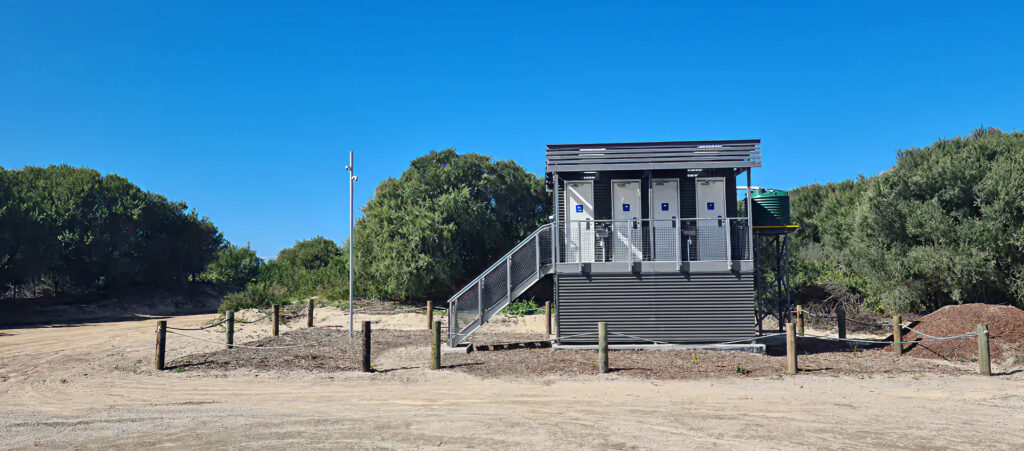 Front view—Belmont Wetlands beachside toilets: raised 4-cubicle building with stairs; composting units beneath; side water tank on stand.