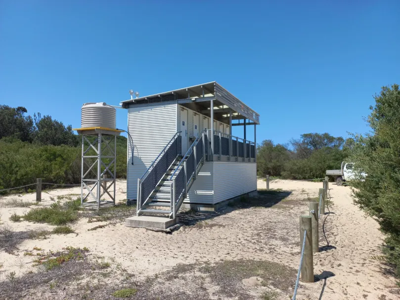 Belmont Wetlands beachside toilets—raised 4-cubicle building with stairs; composting units beneath; side water tank on a stand.