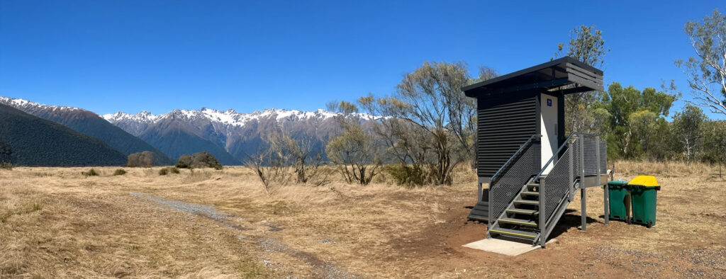Eco-tourism toilet—single cubicle with stair access; composting chamber beneath; snow-peaked mountains and valley backdrop.