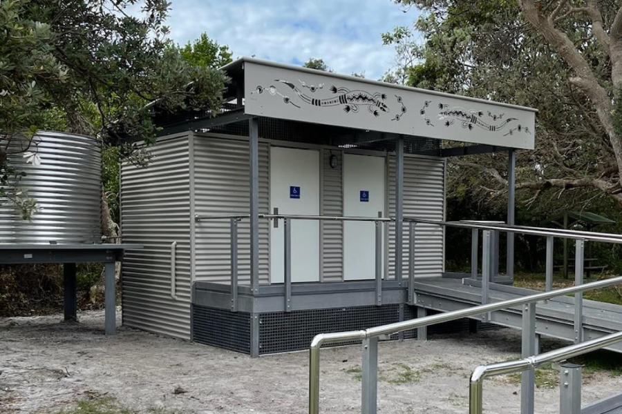 Prefabricated restroom in bush with two accessible cubicles, ramped access, and a side water tank on a stand.